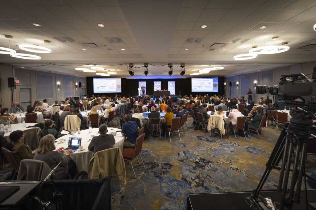 Wide view of the full PEIP conference ballroom, audience seated at round tables facing the stage and presentation screens