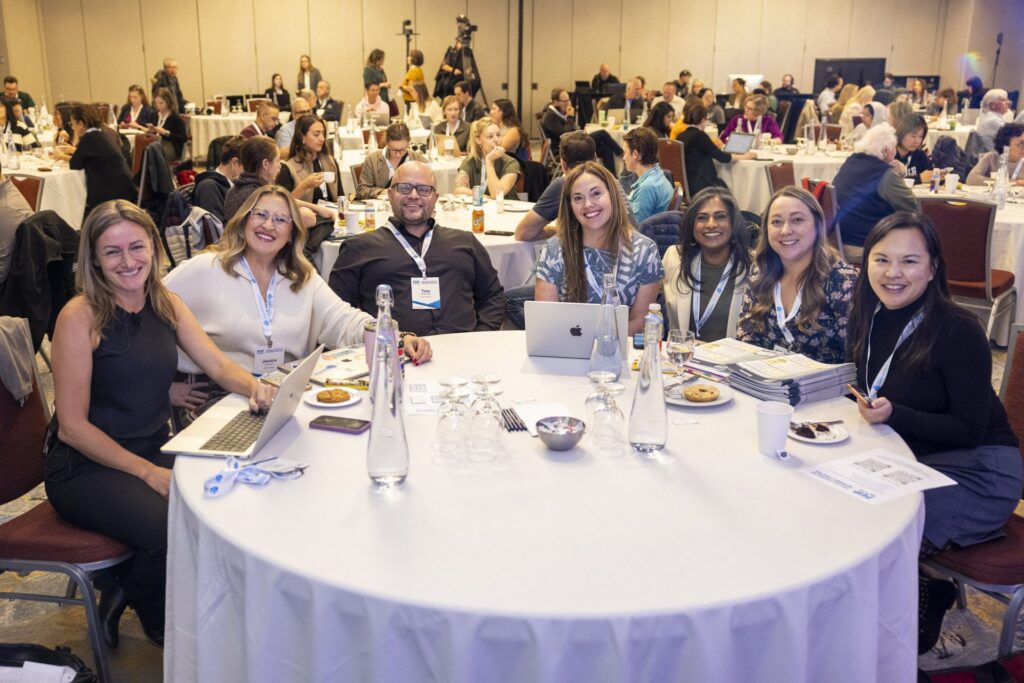 Seven attendees smile around a round table in the conference ballroom, laptops and notes in front of them