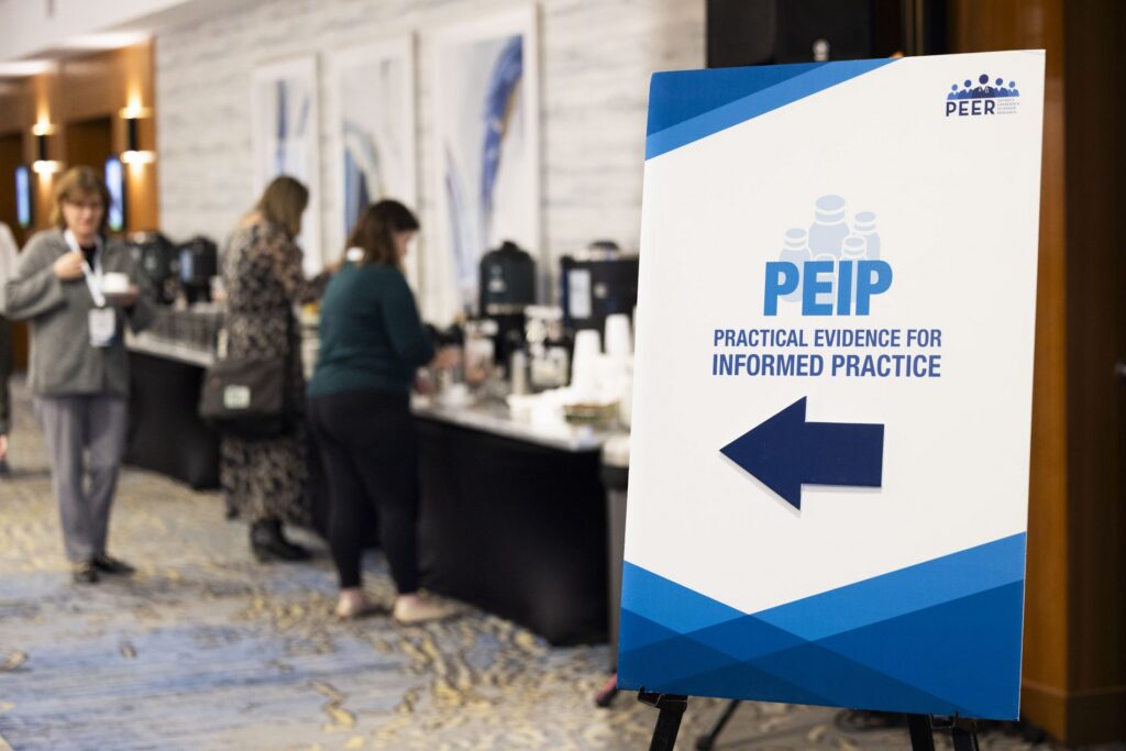 A blue PEIP directional sign with an arrow stands beside the coffee and refreshment station as attendees serve themselves