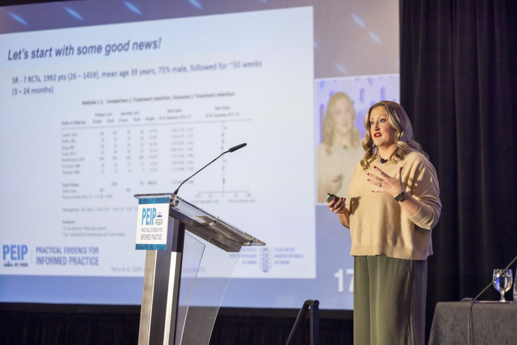 A presenter gestures at the lectern beside a slide showing a data table